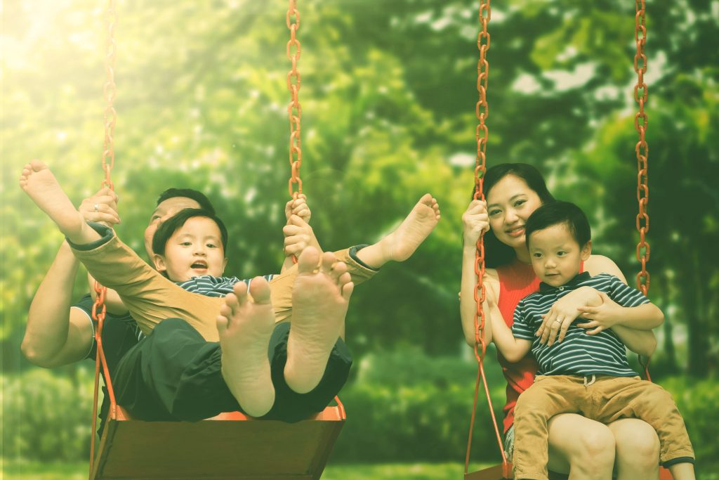 Image of happy parents playing on the swing with their sons. Shot in the park