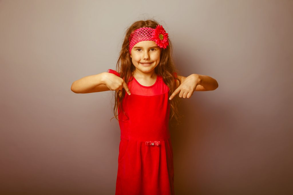 Girl European appearance haired child of seven in red bright dress shows thumb down on the gray background, direction retro