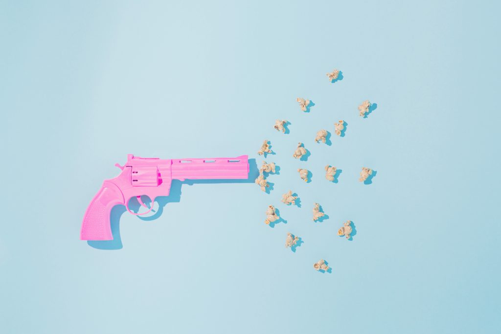 Pink pistol shoots popcorns on pastel blue background. Candy colors. Minimal flat lay concept of non-violence and peaceful conflict resolution.